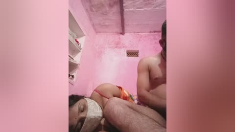 A video captures a young woman with medium skin tone, wearing a white lace bra, lying on a bed in a pink room. Her face is covered by a cloth, and a man is partially visible in the background.