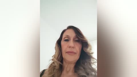 Video of a middle-aged woman with wavy, shoulder-length blonde hair, wearing a beige jacket. She has a neutral expression, looking slightly down, with a soft, diffused background.