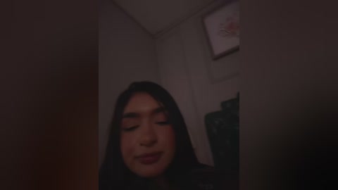Video of a young woman with long black hair, closed eyes, and a slight smile, standing in a dimly lit room with a framed picture on the wall.