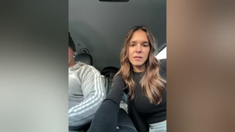Video of a young woman with long, wavy blonde hair, wearing a black sweater, sitting in a car. The car's interior is dimly lit, and a man in a striped jacket is visible in the background.