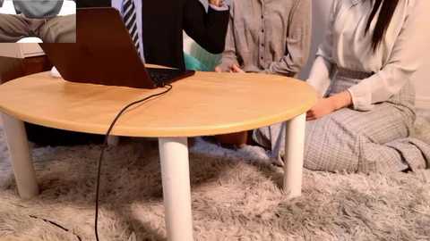 Video of a modern office scene featuring a light-wooden table with white legs, occupied by a laptop and two people, one seated and one standing. The background includes a white shag rug and office furniture.