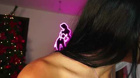 Video of a woman with long black hair, adorned with a neon pink light-up figure of a woman in a bikini, standing in a softly lit room with a Christmas tree and red decorations.