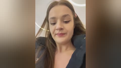 A close-up video of a young woman with long, straight brown hair, wearing a black jacket. She has light skin and a slight smile. The background is blurred, focusing attention on her face.