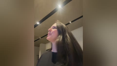 Video of a woman with long, straight brown hair, wearing a black dress, standing in a modern, dimly lit room with recessed lights on a high ceiling.
