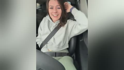 Video of a smiling Caucasian woman with shoulder-length brown hair, wearing a light grey jacket and grey seatbelt, sitting in a car.