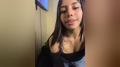 A video of a young Latina woman with medium skin tone and long, straight black hair, wearing a black top with a low neckline, standing in a beige-walled room with a TV visible.