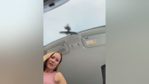 A video of a blonde woman in a pink tank top, standing in a modern, minimalist bathroom with a white ceiling, gray walls, and a visible showerhead. The image is slightly blurred, emphasizing a sense of movement.