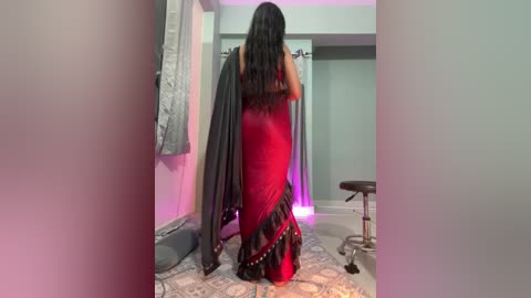 Video of a woman in a red saree, with a grey shawl, standing in a dimly lit room with light blue walls, a grey curtain, and a stool.