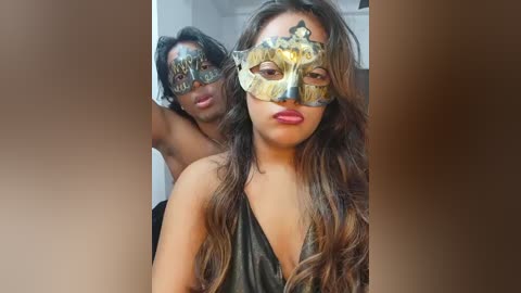 Video of two women in black leather outfits, wearing elaborate golden Venetian masks with intricate designs, set against a blurred, beige-toned background.