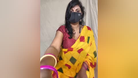 Media: A video of a South Asian woman with medium-dark skin, black hair, and a black mask, wearing a yellow sari with red and green diamond patterns, holding a pink bracelet.