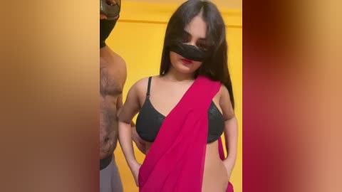Video of a topless, dark-skinned man wearing a red sari and black mask, standing in a yellow room with a hand on his hip, looking confident.