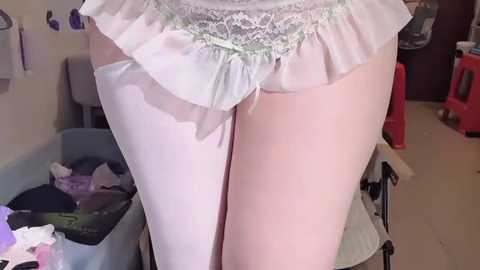 Video of a fair-skinned woman's legs and buttocks in sheer white stockings, partially visible white lace panties, standing in a cluttered laundry room with laundry baskets and a red chair in the background.