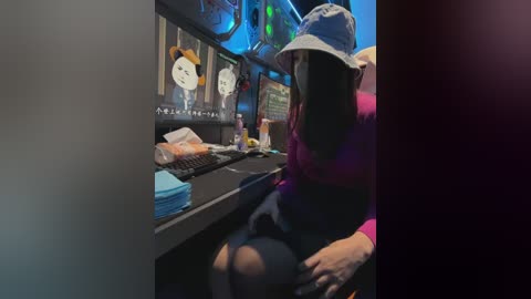 Video of a woman with long dark hair in a light blue bucket hat, wearing a pink long-sleeve shirt and black skirt, sitting at a computer desk with multiple screens displaying cartoon characters, cluttered with papers and drinks.