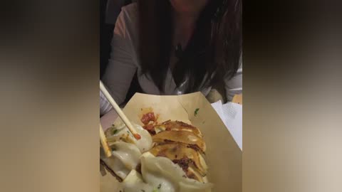 Video of a woman with long dark hair, eating a jiaozi (Chinese dumpling) with a spoon, from a slightly elevated angle, showing a close-up of the dumplings in a brown paper container.
