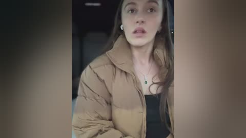 Video of a young woman with fair skin and long, dark hair, wearing a tan puffer jacket over a black top, looking surprised, taken indoors with blurred background.