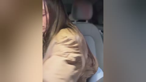 Video of a woman with long brown hair, wearing a beige jacket, seated in a car. The image is blurry and cropped, focusing on the side profile.