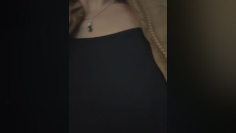 A close-up video of a woman's upper body in a black dress, showing a delicate gold necklace with a small pendant. Her blonde hair is partially visible. The background is dark and indistinct.