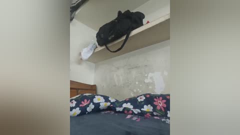 Video of a small, cluttered bedroom with a bed covered in dark bedding with pink and white flowers, a black backpack hanging from a shelf, and peeling white walls.
