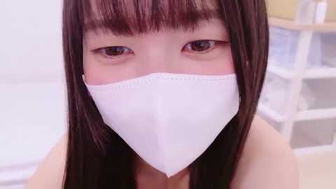 Video of an Asian woman with long, straight black hair, wearing a white surgical mask, and brown eyes. Background shows a white shelf with books and a beige wall.