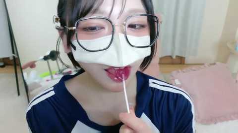 Video of an Asian woman with glasses and a white face mask eating a lollipop, wearing a navy blue shirt with white stripes. The background shows a room with light walls, a white chair, and a pink pillow.