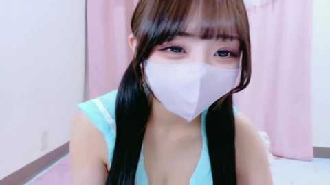 Media: A video of a young Asian woman with straight, long black hair and pale skin, wearing a light blue tank top and a white face mask, standing in a room with pink curtains and white walls.