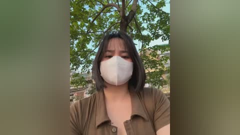 Video of an Asian woman with shoulder-length black hair, wearing a face mask, brown blouse, and greenery background.