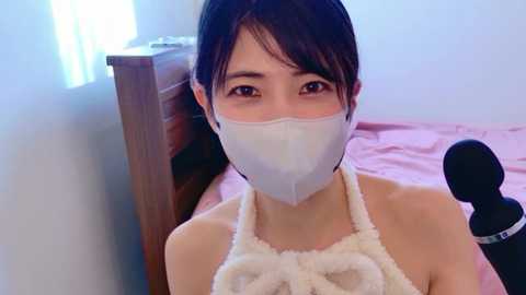 Video of a young East Asian woman with short dark hair, wearing a white face mask and a white halter top, sitting on a pink bed with a black vibrator. The room is brightly lit with natural light.