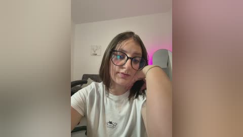 Video of a young woman with shoulder-length brown hair, wearing black-framed glasses and a white T-shirt, leaning against a wall in a dimly lit room with pink and purple lighting.
