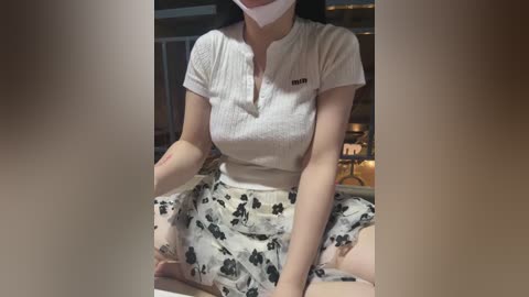 Video of a fair-skinned woman with a light complexion, wearing a white ribbed tank top and a black floral skirt, seated cross-legged in a dimly lit room with a blurred background.