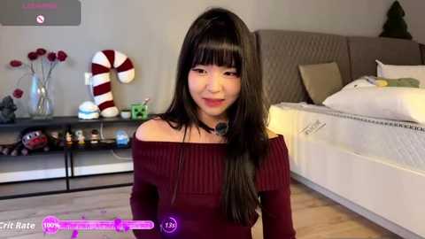 Video of an Asian woman with long black hair and bangs, wearing a burgundy off-shoulder sweater, in a modern bedroom with a beige bed, white pillow, and Christmas decor.