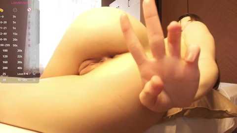 A close-up video of a nude person with fair skin, lying on a bed, making a peace sign with their hand. The background includes a TV screen displaying a calendar and a wooden cabinet.