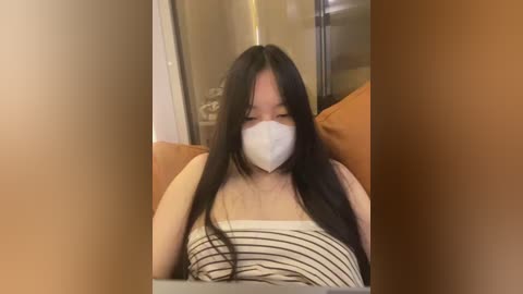 Video of an East Asian woman with long black hair, wearing a white face mask, striped tank top, and orange pillow, indoors, looking down.