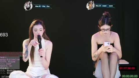 Video of two Asian women on stage, one in a white dress, the other in a black strapless dress, singing into microphones, with a digital scoreboard in the background.