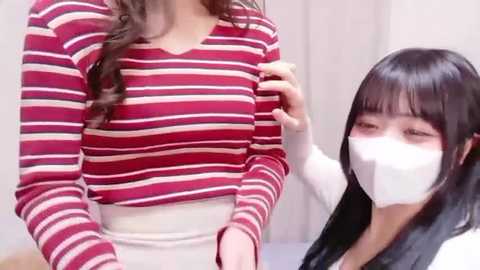 Video of an Asian woman with long black hair, wearing a white mask, adjusting the red and white striped top of another woman, who is partially visible.