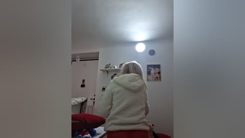 Video of a blonde woman in a light green hoodie sitting at a red stool in a small, dimly-lit room with white walls, a clock, and framed pictures.