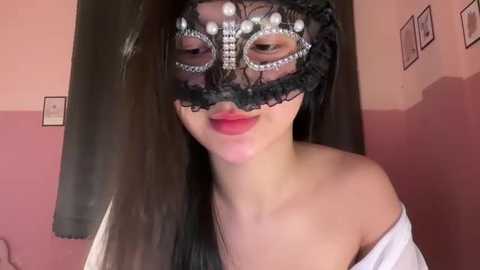 Video of a woman with long black hair, wearing a black lace masquerade mask adorned with pearls, and a white off-shoulder dress.