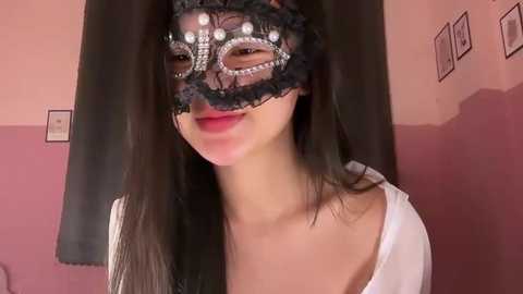 Video of an Asian woman with long black hair, wearing a black lace masquerade mask adorned with silver studs, a white top, and a pink and white two-tone wall background.