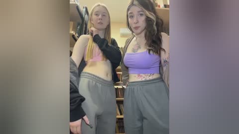 Video of two young women with light skin, one with long blonde hair, the other with long brown hair, both in casual outfits, standing in a cluttered room with books and shelves in the background.