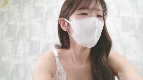 Media: A video of an Asian woman with long dark hair, wearing a white face mask, white lace top, and a light-colored background.