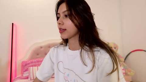 Media: A young, light-skinned Latina woman with long, dark brown hair, wearing a white T-shirt with a cartoon character, stands in a cozy bedroom with pink decor, a bed with a tufted headboard, and a red lamp.