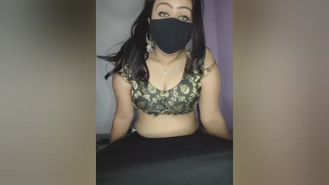 Video of a South Asian woman with medium brown skin, wearing a black mask, green floral crop top, and black pants, seated against a purple backdrop.