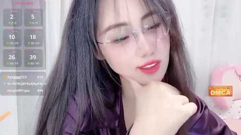 A video of a young Asian woman with long, straight black hair, fair skin, and clear glasses. She has full lips painted in bright red lipstick. She is wearing a dark purple satin robe.