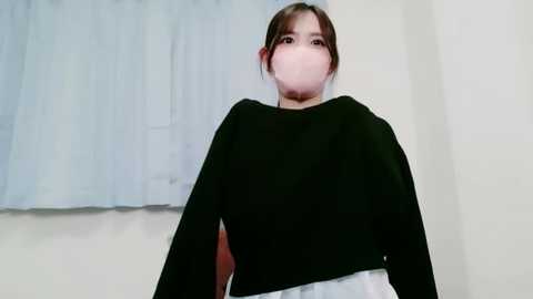 A video of a young East Asian woman with a fair complexion, wearing a black long-sleeved top and white skirt, standing indoors near a white curtain. She has a surgical mask covering her mouth.