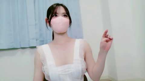 Video of an East Asian woman with pale skin and straight, dark hair tied back, wearing a white lace dress and pink face mask, gesturing with her right hand, standing indoors against a light blue curtain and white wall.