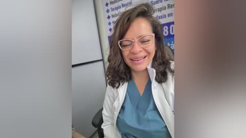 Video of a smiling, middle-aged Caucasian woman with wavy brown hair, wearing glasses and a white lab coat over a blue scrub top, seated at a desk with medical posters in the background.