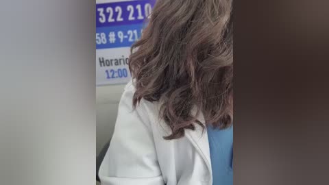 Media: Video of a woman with wavy, shoulder-length brown hair, wearing a white lab coat, standing in a medical office with a blue sign and phone number on the wall.