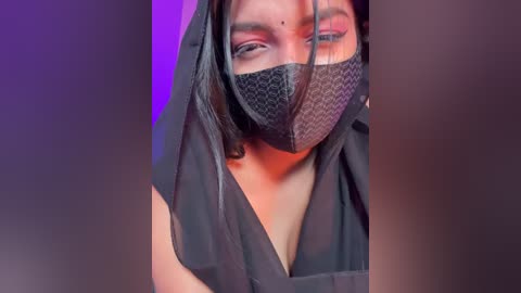Video of a South Asian woman with medium skin tone, wearing a black mask and black headscarf, with a faint purple and red gradient background.