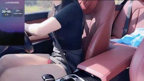 Video of a woman with long black hair, wearing a black top and gray shorts, driving a car with red leather seats, sunlight streaming in.