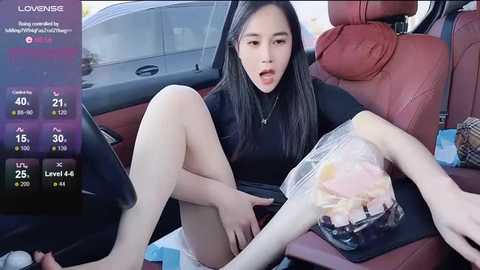 A candid video shows an Asian woman with long black hair, wearing a black dress, sitting in a car with a plastic bag beside her.
