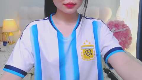 Video of a young woman with fair skin wearing a blue and white Argentina soccer jersey, red lipstick, and a pink hair accessory. Background features a bed with a floral duvet, a lamp, and a pink crystal.